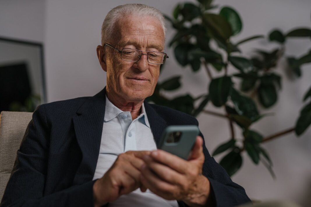 Starszy mężczyzna w okularach uśmiecha się, korzystając ze smartfona w domu Starszy mężczyzna w okularach uśmiecha się, korzystając ze smartfona w domu